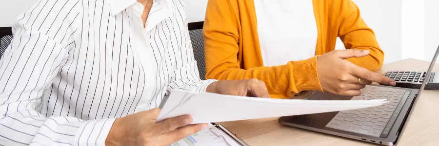 Dos personas sentadas en una mesa, una sosteniendo un documento y la otra se帽alando la pantalla de una laptop, enfoc谩ndose en una tarea colaborativa.