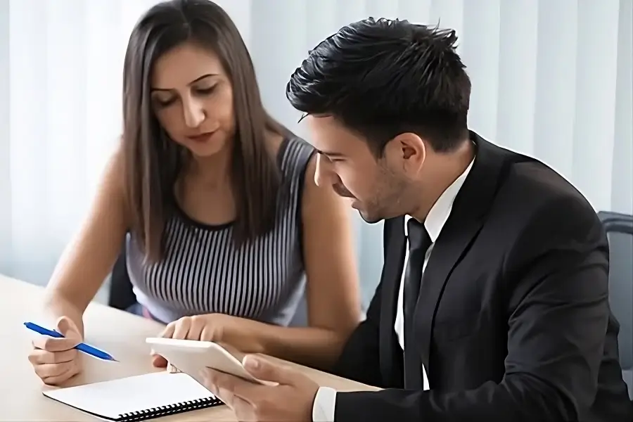 Un hombre y una mujer sentados en una mesa, el hombre sostiene una tableta y parece estar explicando algo a la mujer que tiene un cuaderno frente a ella.